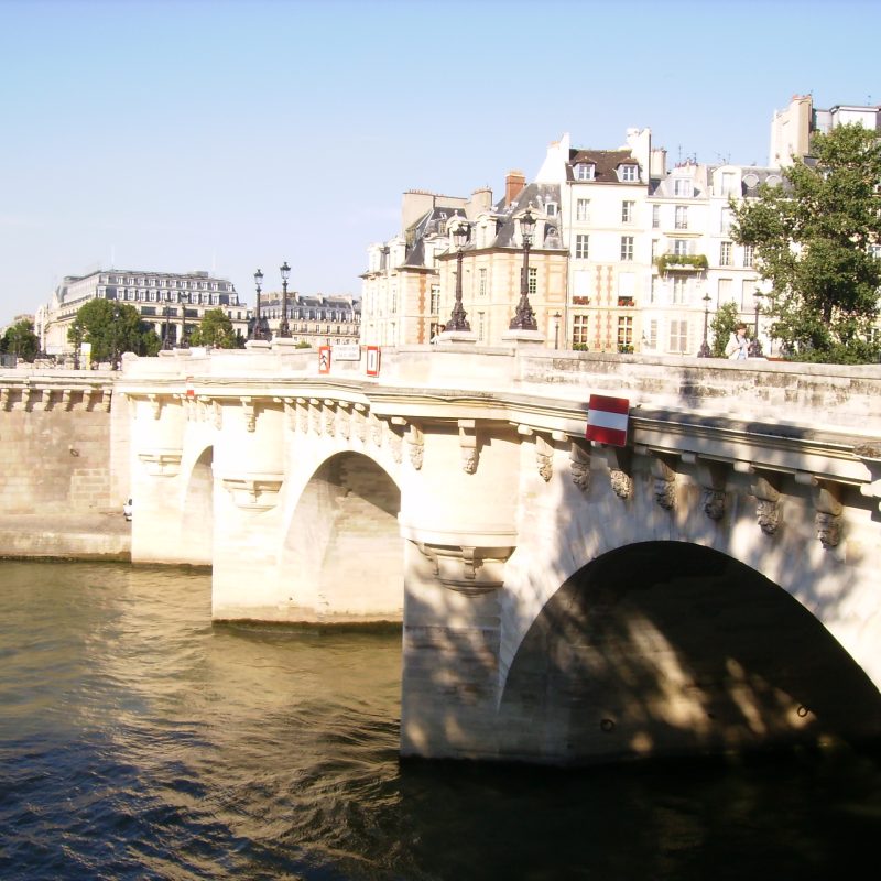 Vue du Pont Neuf à Paris par une journée ensoleillée d'été, avec des gens profitant de promenades le long de la Seine et des bateaux naviguant sur le fleuve