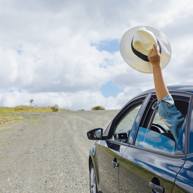 A person sticking their arm out of a car window with a hat, symbolizing budget travel