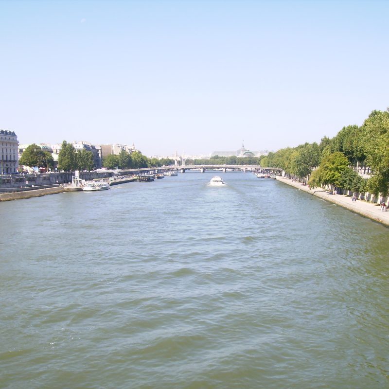 Vista del Río Sena en París en un día soleado de verano, con gente disfrutando de paseos por la orilla y embarcaciones navegando por el río