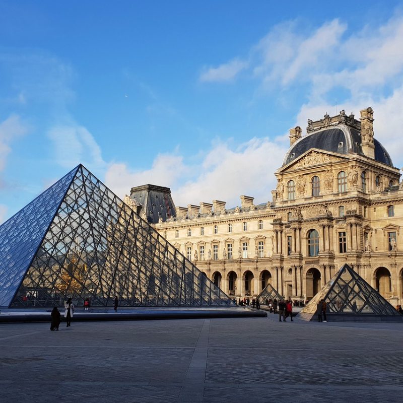 View of the Louvre Plaza in Paris on a sunny summer day, with people enjoying the outdoor space and the iconic Louvre Pyramid in the background