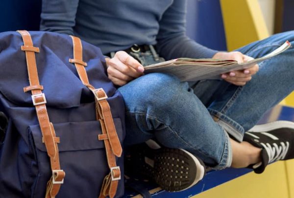 Personne assise sur un banc avec un sac à dos à côté, lisant un journal