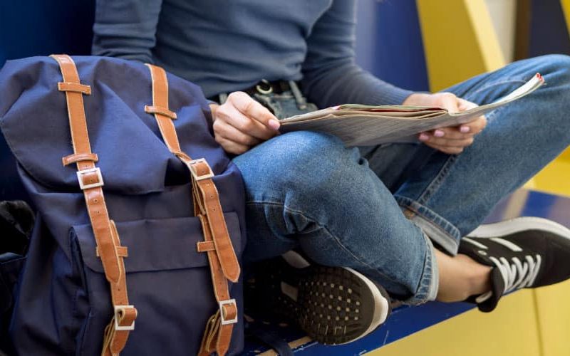 Personne assise sur un banc avec un sac à dos à côté, lisant un journal